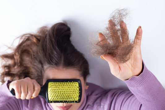A woman laying on the surface holding a hair brush over her face with one hand and holding hair of clumps in another 