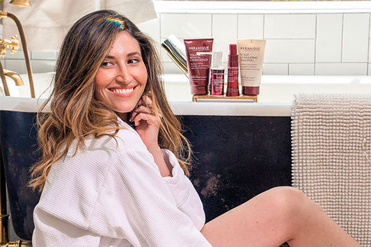 A woman sitting on the bathroom floor in front of the tub with keranique products placed on the edge of the tub